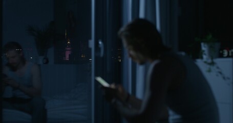 A man in pajama pants uses a mobile phone while sitting on the bed at night. A blurred silhouette. Dark room, home interior