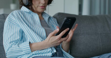 A woman in casual clothes and with headphones is using a mobile phone and smiling. She's sitting on the couch at home. Daylight