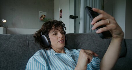 A woman with headphones on her head uses a mobile phone while sitting on the couch at home. She smiles and dances with pleasure. Daylight