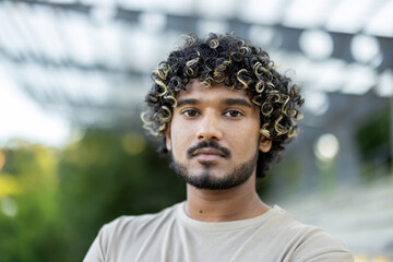 Close-up portrait of a young calm and serious man, looking at the camera, walking outside in the park.