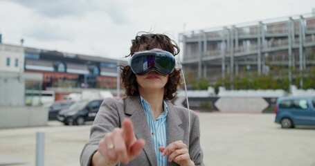A stylish woman in a jacket wearing a virtual reality headset stands on a city street and moves her arms. Summer day