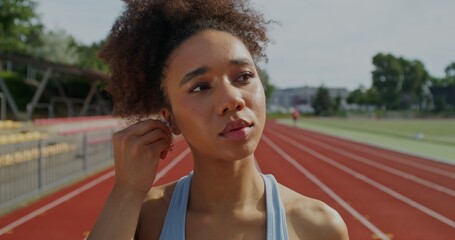 A woman in sportswear puts on earphones while standing at the city stadium. She's getting ready for...