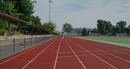 A woman in sports clothes and with earphones in her ears runs at the stadium on a summer day