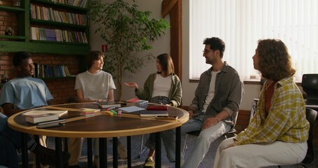 A group of young people are smiling and talking while sitting around a round table indoors. Notebooks and books on the table. Daylight
