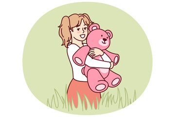 Happy little girl with teddy bear in hands stands in meadow among tall grass and looks at screen. Child with smile hugs beloved toy bear, presented for birthday, during summer walk