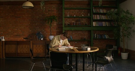 A man is studying the Bible while sitting at a table indoors. He writes in a notebook and leaves notes on stickers. Daylight