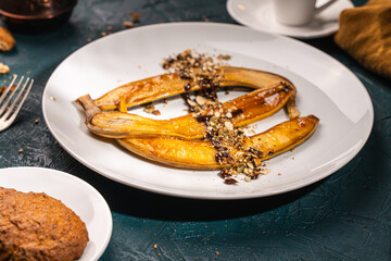 Bananas flambe on a white plate with nuts and chocolate on a table with coffee and a cezve on a dark background with a knife and fork. Restaurant serving dessert