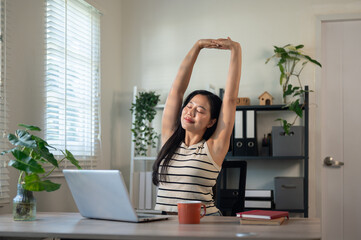 A carefree Asian businesswoman is stretching her arms to relax after finishing her work on a laptop.