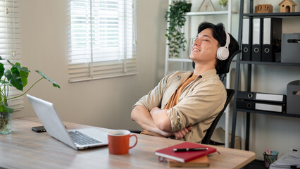 A relaxed Asian businessman is leaning back in his chair, enjoying the music on his headphones.