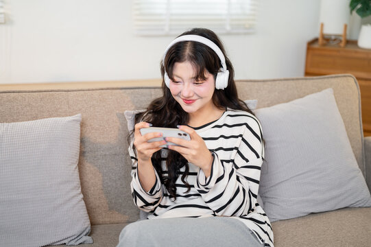 A happy Asian woman wearing headphones enjoys watching a movie on her smartphone on a sofa.