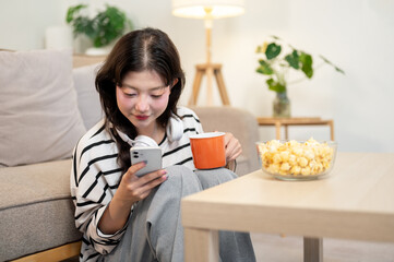 A beautiful, relaxed Asian woman is sipping coffee and reading chats on her smartphone at home.