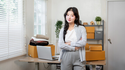 A confident Asian female online business entrepreneur stands with her arms crossed in her office.