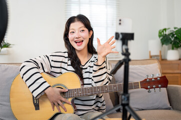 Cheerful Asian female influencer is performing on the guitar in her living room on her social media.