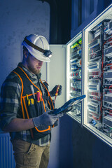 Electrician in overalls, focused on work in switchboard with fuses, using tablet.