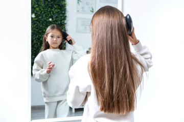 Fototapeta premium Little girl combing her long brown hair in front of the mirror