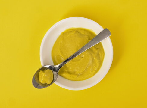 Mustard in ceramic cup with spoon on yellow background.