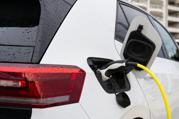Fototapeta premium Close up view of white electric car charging via charging plug pistol on urban city street. Charging process as concept of sustainable living, eco-friendly lifestyle, energy efficient transportation