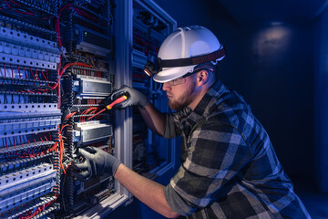 A male electrician works in a switchboard in overalls against the backdrop of emergency lighting.