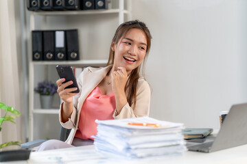 Joyful Businesswoman: A young, smiling businesswoman checks her smartphone, surrounded by a large stack of paperwork in her modern office.  She exudes success and happiness. 