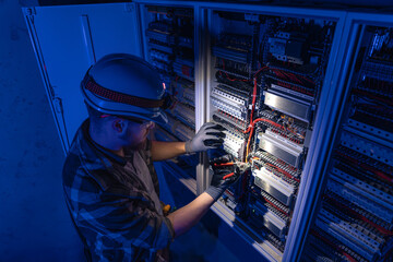 A male electrician works in a switchboard in overalls against the backdrop of emergency lighting.