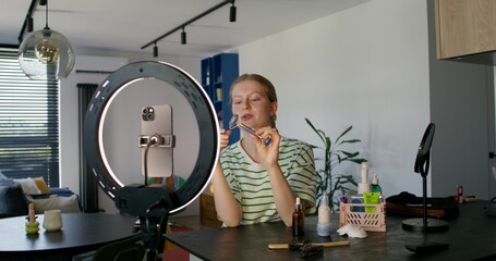 A young woman is doing a facial massage using a roller massager. She smiles and talks while filming a beauty blog on her mobile phone. A smartphone and a round lamp on a tripod