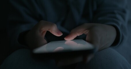 A woman uses a mobile phone while sitting in a dark room. Close-up of her hands, an unrecognizable...