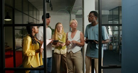 A group of young people of different nationalities are talking standing near a glass board with notes. Business planning. Daylight