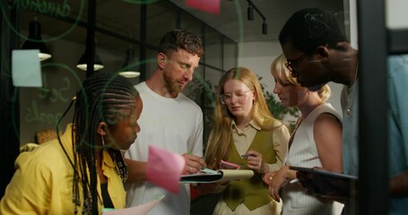 A group of young people are talking standing near a glass board with notes. They stick stickers on the board. Business planning