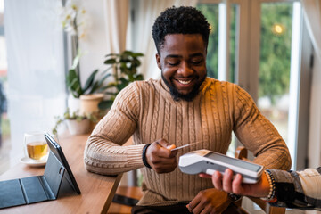 Young man in casual clothing makes a contactless payment with payment card while sitting in the cozy city cafe.