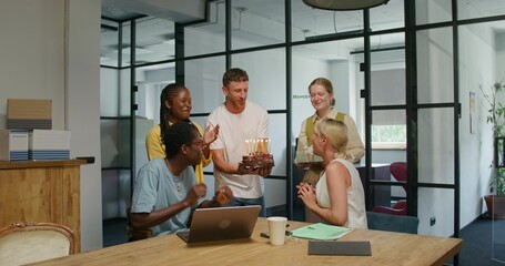 Obraz premium Colleagues wish a woman a happy birthday in the office. She blows out the candles on the cake. Daylight
