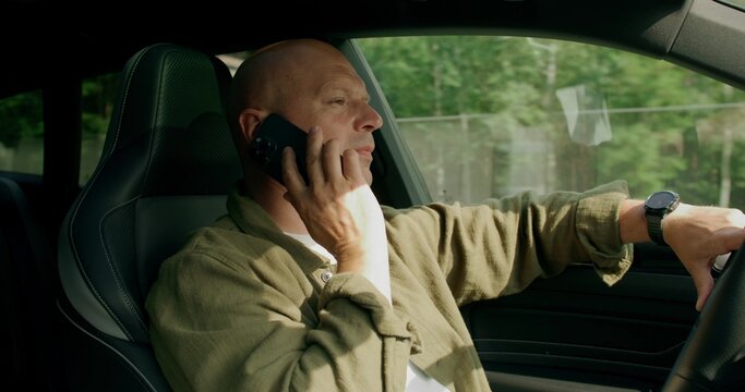 A man is talking on a mobile phone with a serious look while driving a car. View from the car interior. Summer day