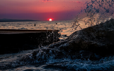 Waves crash dramatically against rocks as the sun sets over the horizon, casting vibrant red and...