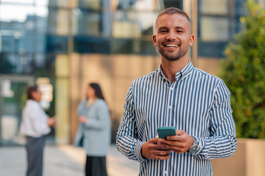 Young manager using smartphone smiling at camera in front of office building