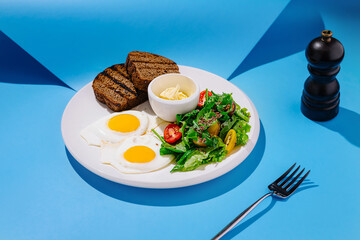 Delicious breakfast featuring sunny side up eggs, dark bread, fresh salad, and butter on a vibrant blue background