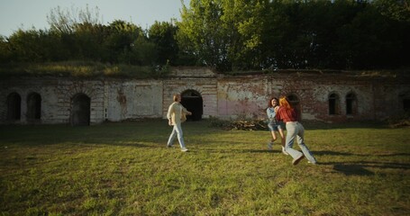 Three young friends are having fun and running around the ruins of a historic building. The fortress walls are overgrown with green vegetation. A sunny summer day