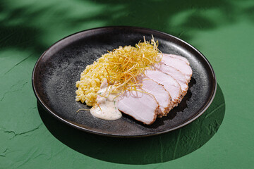 Gourmet dish featuring sliced chicken, savory grains, and sauce on a modern black plate against a vibrant green background