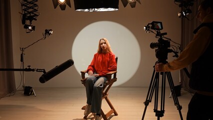 Studio for photo video production filming with lighting equipment and cyclorama, model actress sits in front of camera on chair and talking, recording interview.