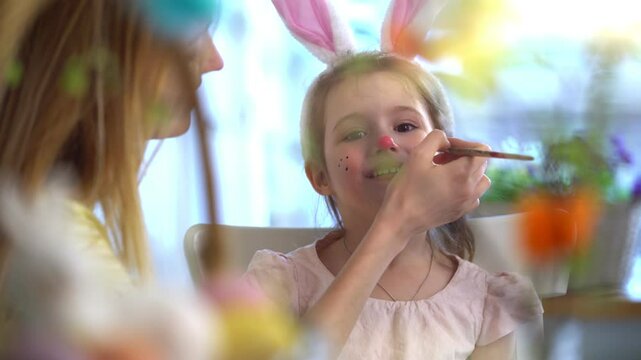 A little girl with bunny ears smiling while getting her face painted by her mother. A joyful Easter moment with warmth, creativity, and family bonding. Soft lighting, colorful decorations, festive and