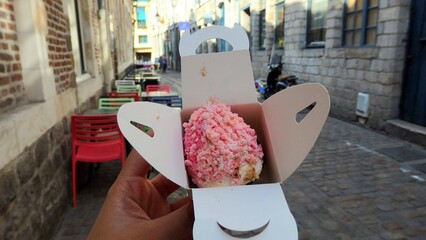 Hand holding Aux Merveilleux de Fred pink cake inside a paper packaging on the small alley outside the shop