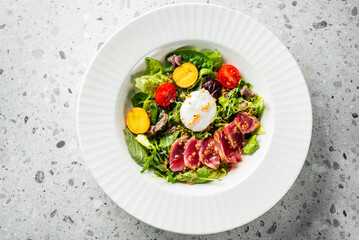 A beautifully arranged salad featuring seared tuna, fresh greens, cherry tomatoes, and roasted potatoes, topped with a poached egg and a sprinkle of nuts, served in a stylish white bowl.