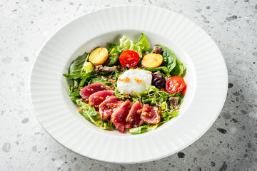 A beautifully arranged salad featuring seared tuna, fresh greens, cherry tomatoes, and roasted potatoes, topped with a poached egg and a sprinkle of nuts, served in a stylish white bowl.
