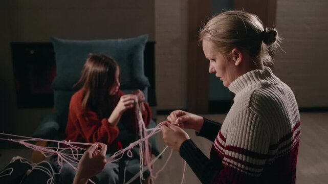  Family members untangle pink yarn together in a cozy room near a glowing fireplace, creating a warm and peaceful bonding moment.
