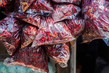 Close-up shot of numerous bags filled with vibrant dried red chilies, hanging at a market. The...