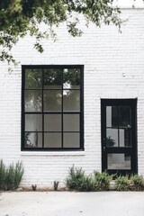 Minimalist architecture showcases a white brick building with large black-framed windows and a sleek black door surrounded by lush greenery during daylight