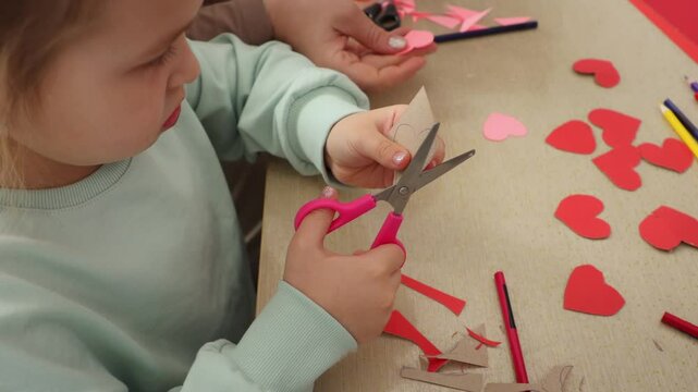 Top view of a little Caucasian girl cutting paper hearts with scissors. Close-up. Concept of St. Valentine's Day and pre-school education.