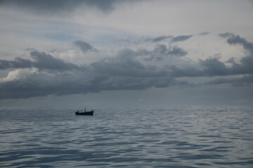 Naklejka premium A solitary boat floats on serene waters, surrounded by a vast expanse of calm ocean and a sky filled with dark, dramatic clouds at twilight.