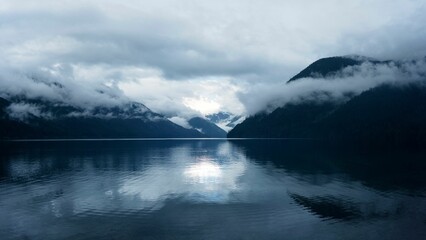 A serene lake with misty mountains reflecting in the water under a cloudy sky.