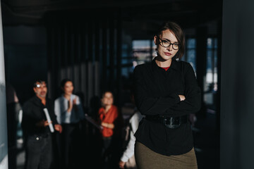 A confident businesswoman stands with her arms crossed, leading a meeting with her team. Colleagues...