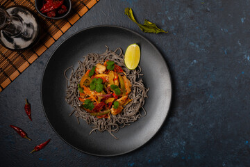 Wheat noodles with black sesame, fried in a wok with chicken and vegetables. Top view. Served on a black plate.