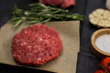 The concept of cooking a burger, fast food, natural products. Close-up of beef raw round patty on paper surrounded by seasoning, herbs and vegetables out of focus on dark background. High quality